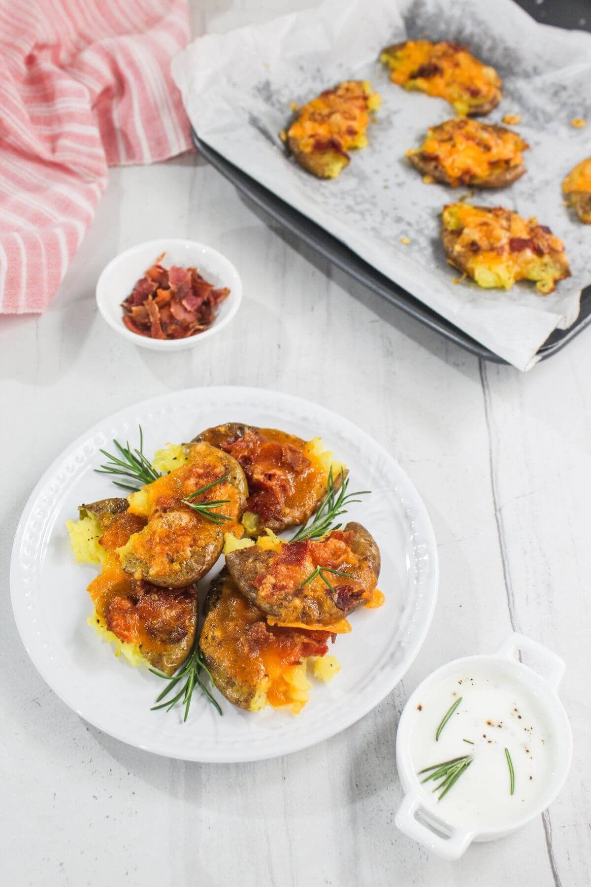A plate of smashed potatoes topped with melted cheese and bacon, garnished with rosemary. Additional potatoes, bacon bits, and a small bowl of sauce are nearby.