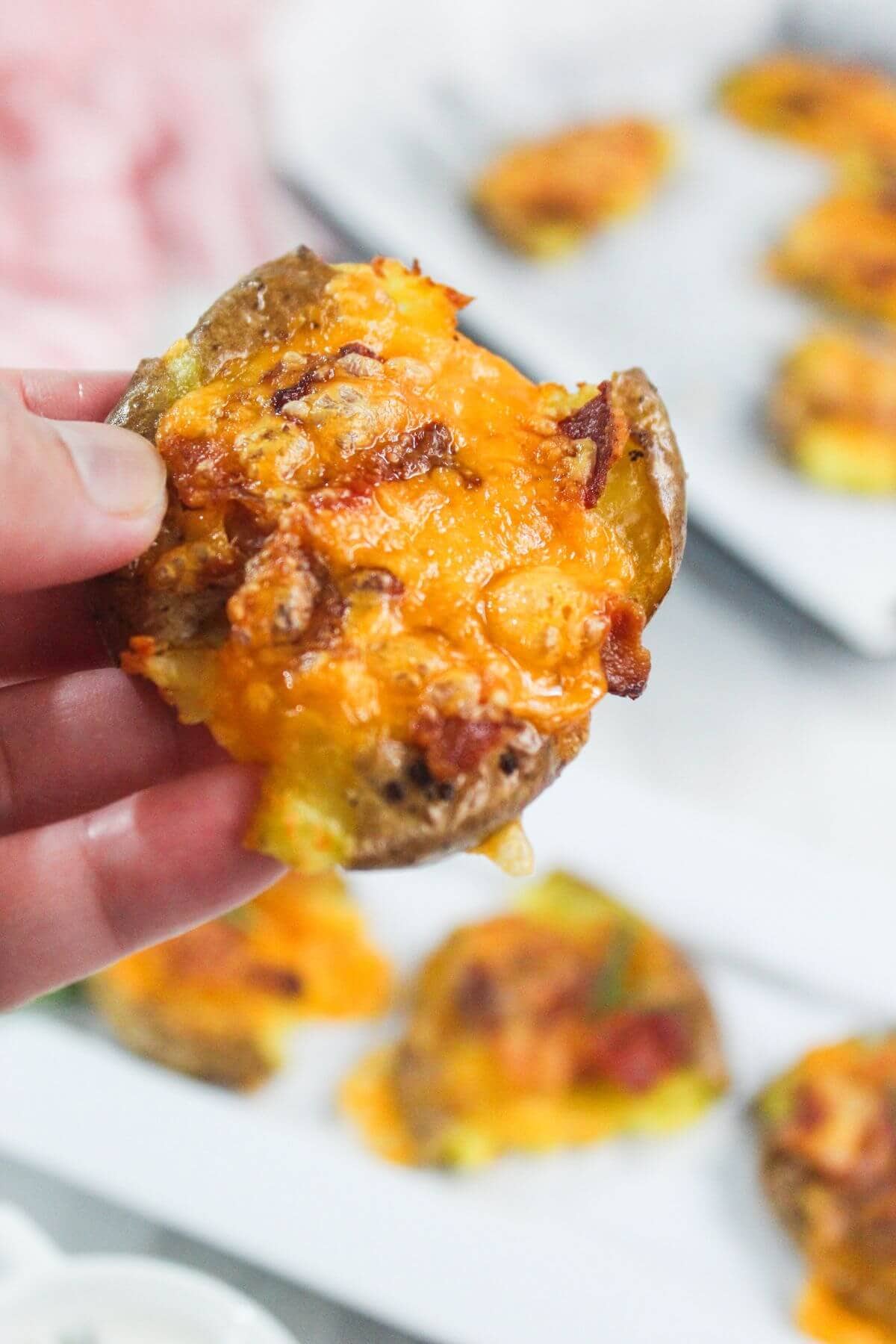 A hand holds a baked potato round topped with melted cheese and bacon bits; more potato rounds are visible on a plate in the background.