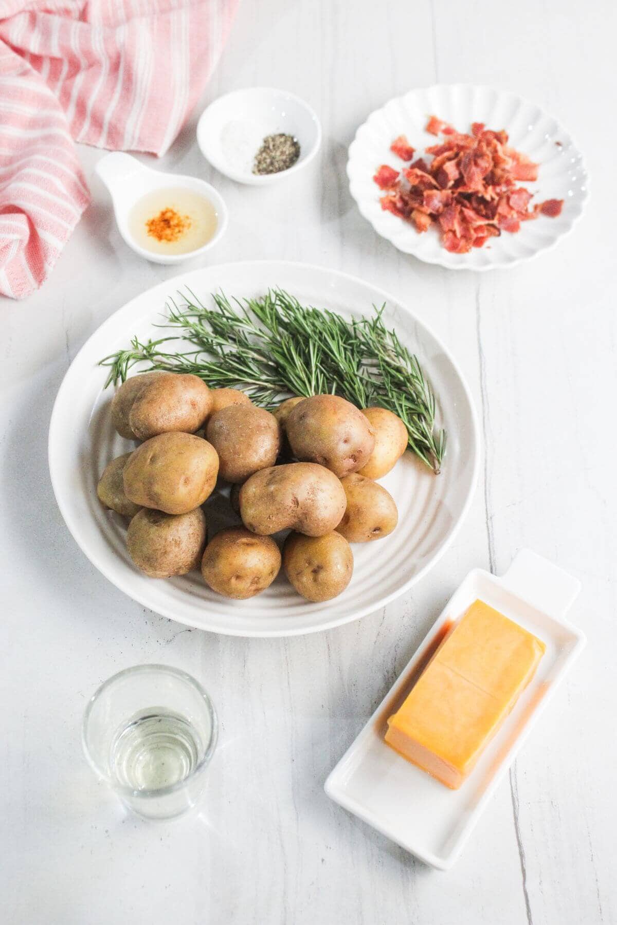 A plate of small potatoes and fresh rosemary, a block of cheddar cheese, a dish of cooked bacon pieces, a bowl of seasoning, and a glass of liquid on a white surface.