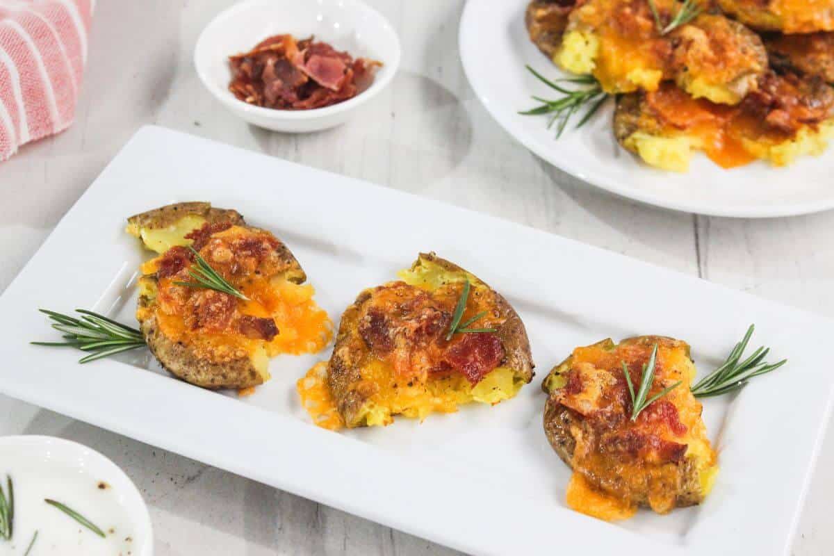 Three smashed potatoes topped with melted cheese and bacon, garnished with rosemary sprigs, are arranged on a rectangular white plate.