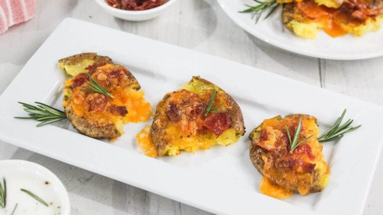 Three baked potato halves topped with melted cheese and bacon bits, garnished with sprigs of rosemary, arranged on a rectangular white plate.