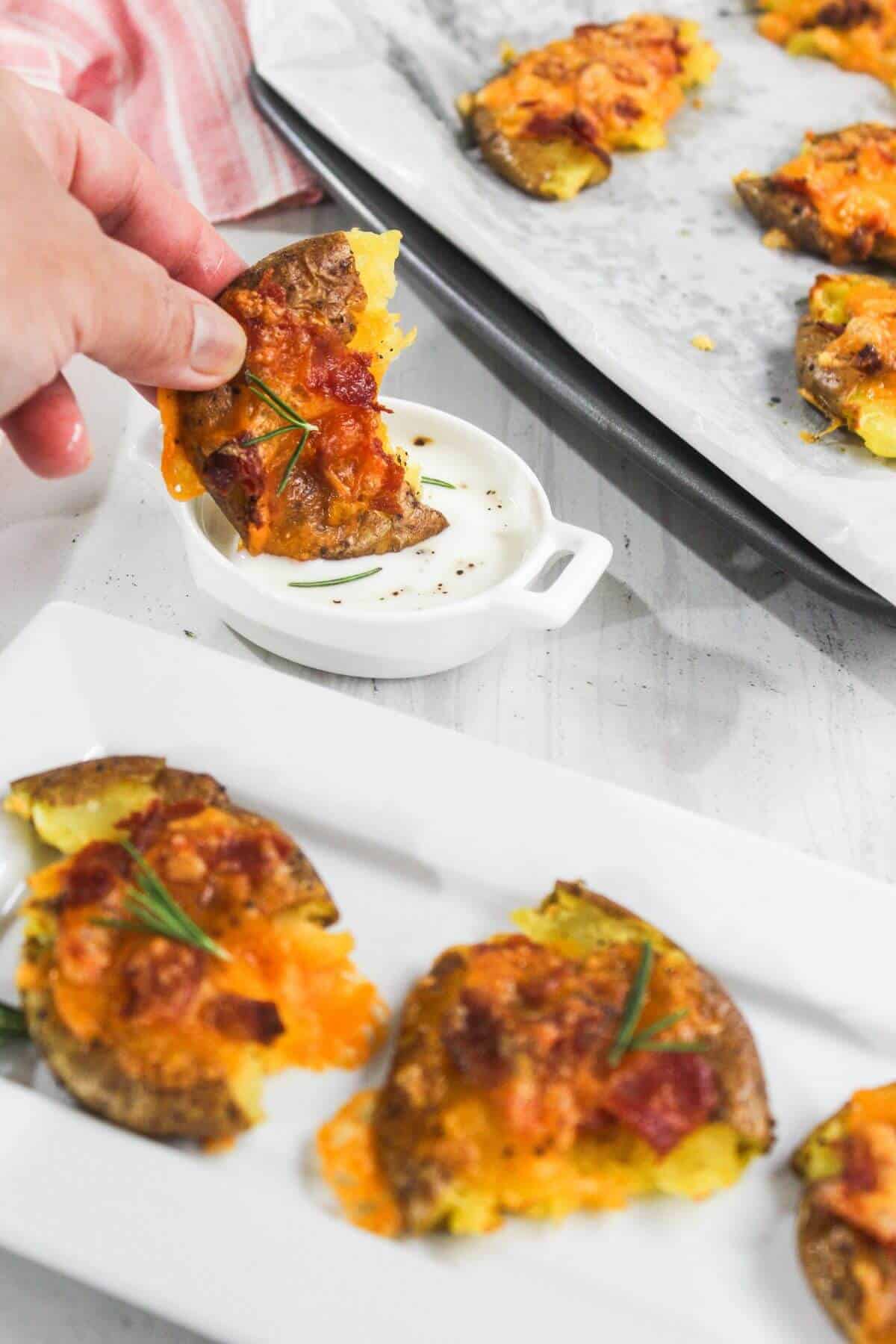 A hand dips a smashed potato topped with cheese and bacon into a small dish of white sauce; more potato pieces are on a plate and a tray.