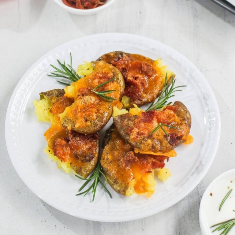 A white plate with four smashed potatoes topped with melted cheddar cheese and bacon bits, garnished with fresh rosemary sprigs.