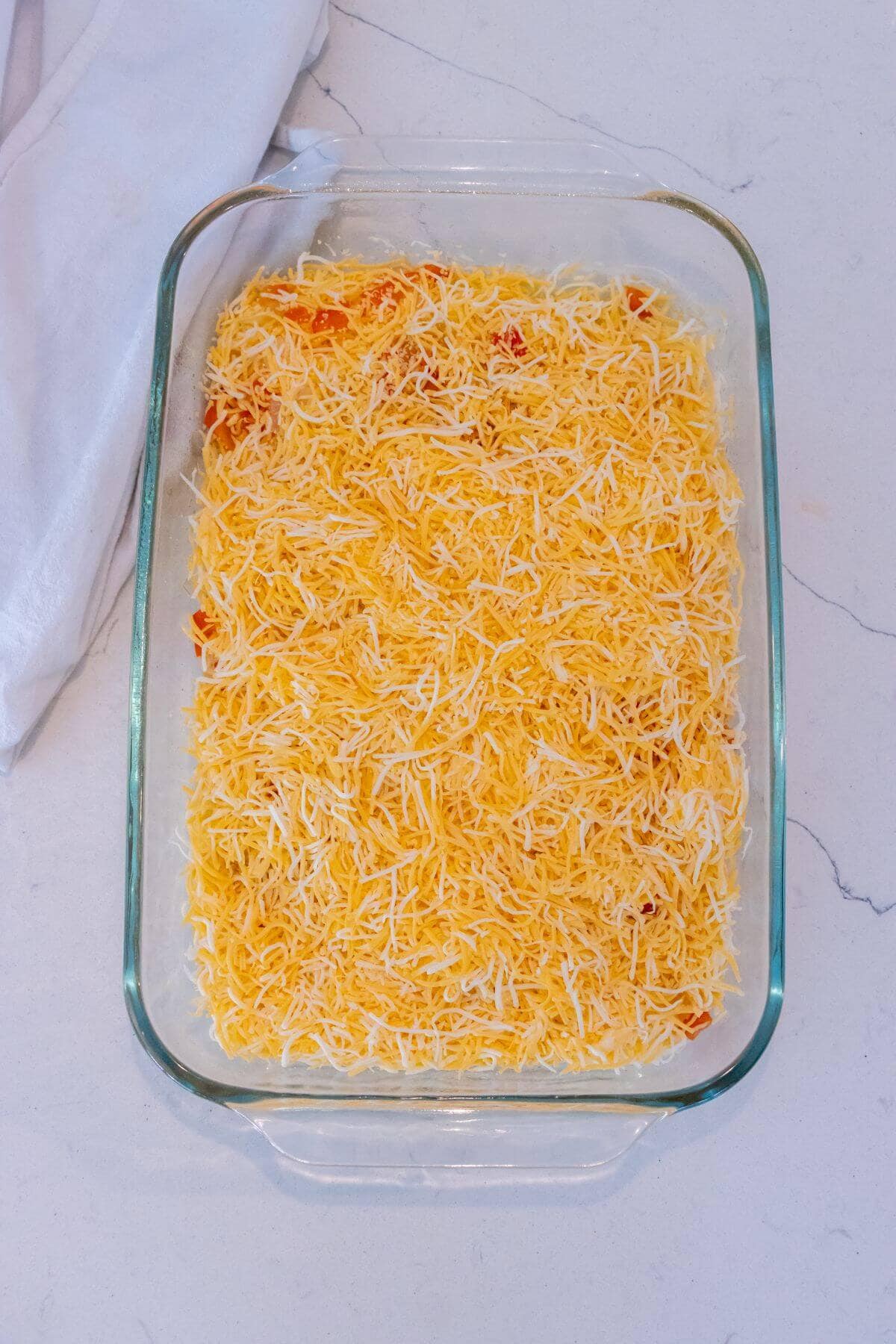 A glass baking dish filled with an even layer of shredded cheese over a mixture, placed on a white countertop with a white cloth nearby.