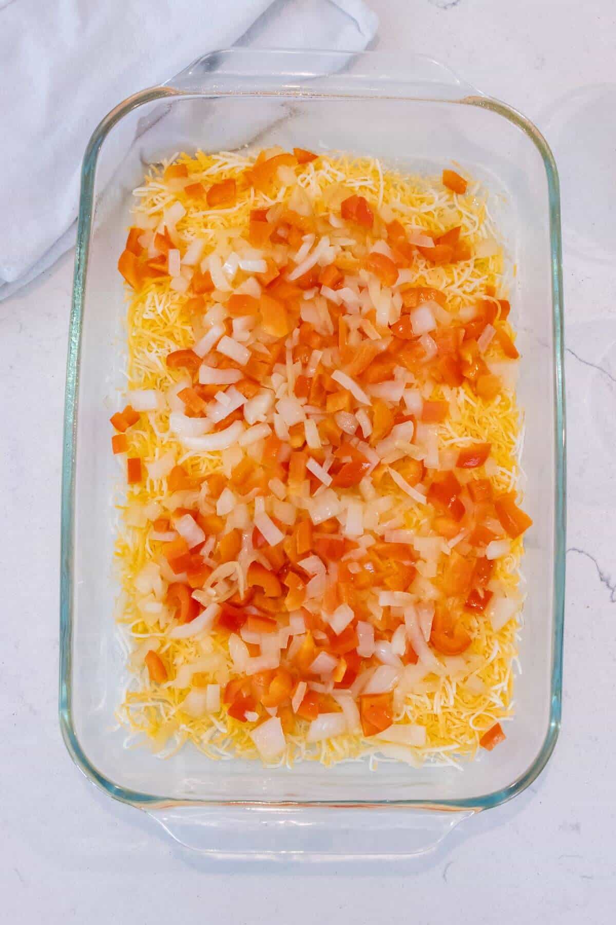 A glass baking dish containing a layer of shredded cheese topped with chopped tomatoes and onions on a white surface.