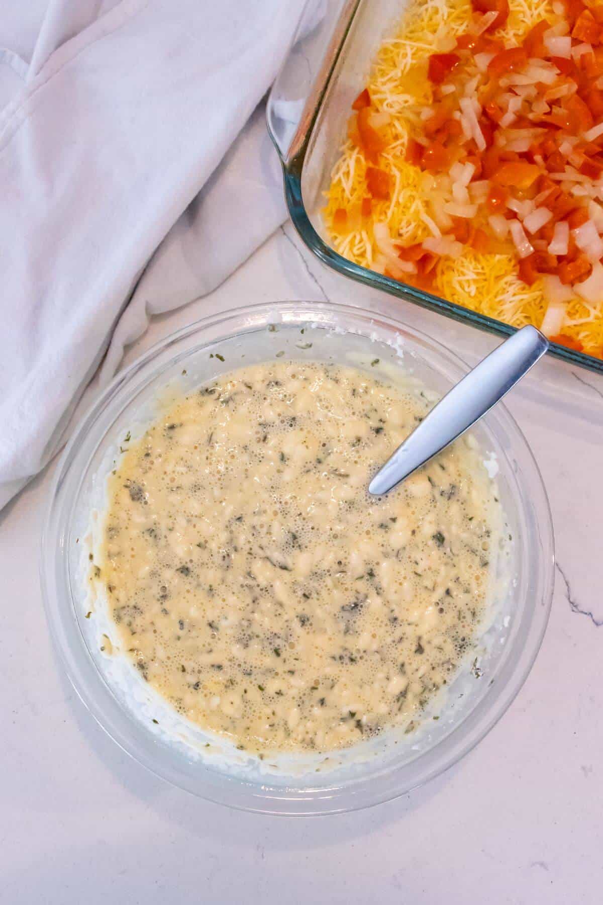 A bowl of creamy herb mixture with a spoon next to a glass dish containing shredded cheese, chopped tomatoes, and onions on a white surface.