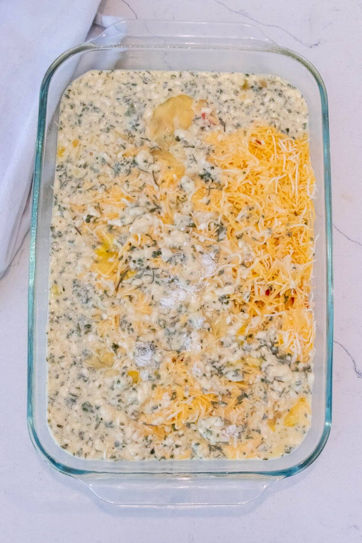 A glass baking dish filled with a creamy spinach and artichoke mixture, partly topped with shredded cheese, ready to be baked.