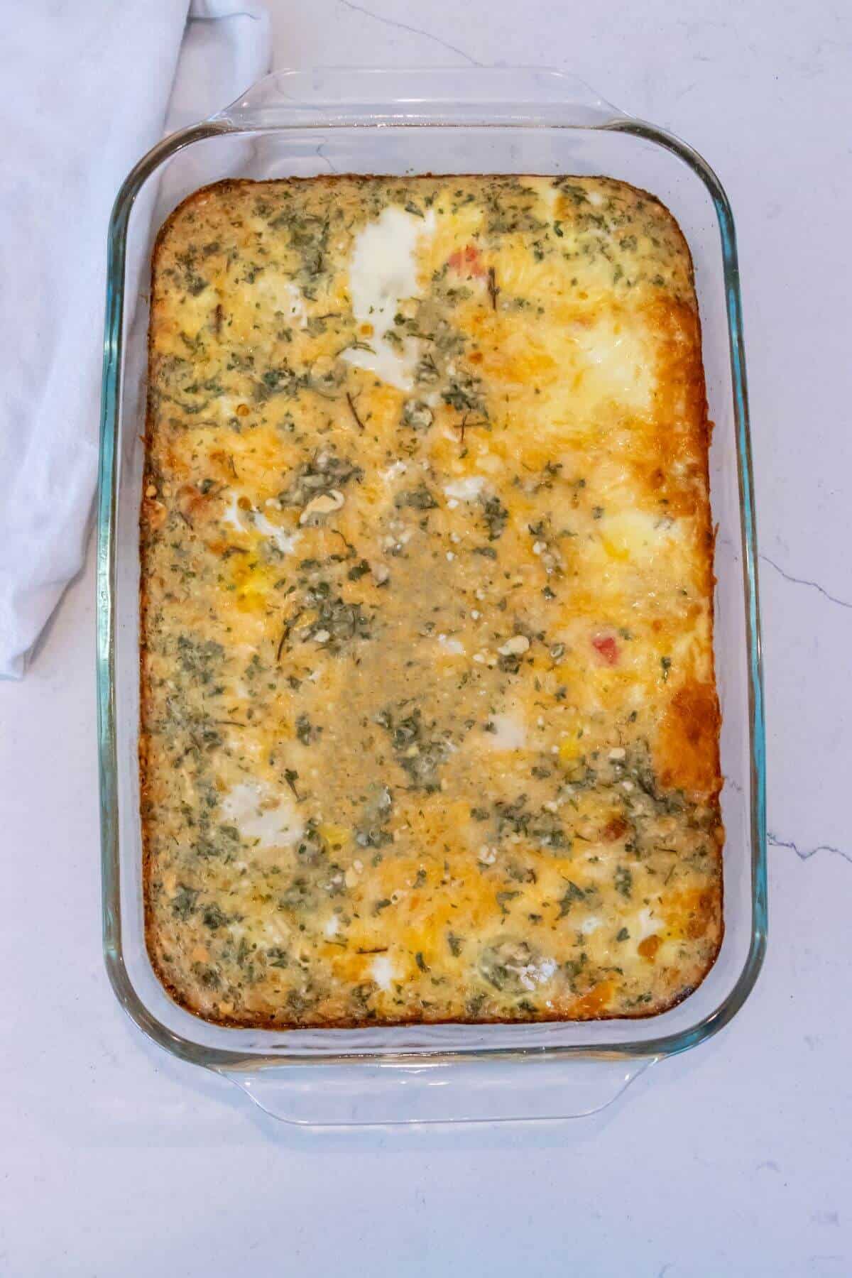 A baked egg casserole in a clear glass dish sits on a light-colored countertop next to a white cloth.