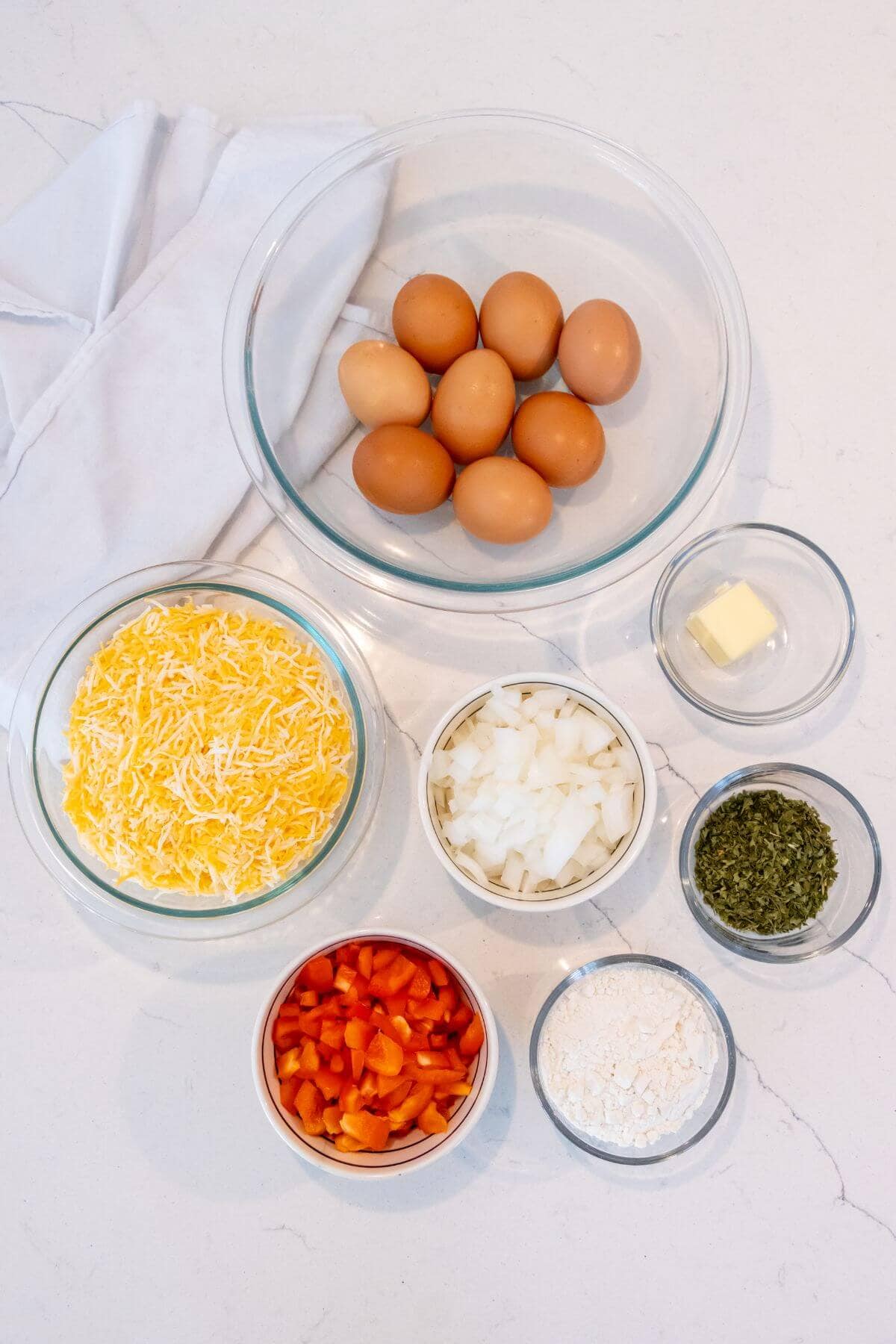 A flat lay of eggs, shredded cheese, diced onion, chopped bell pepper, butter, flour, and dried herbs in glass bowls on a white surface with a folded cloth.