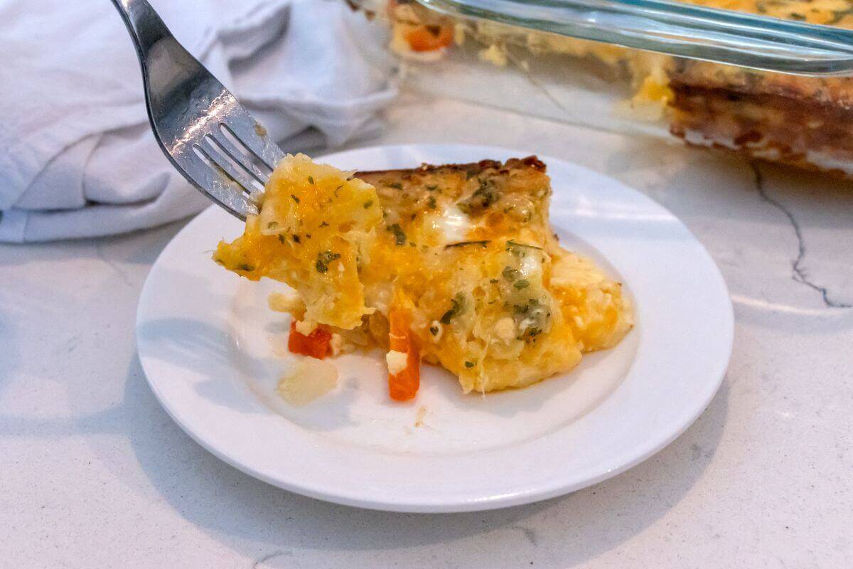 A fork holding a piece of cheesy egg casserole with vegetables on a white plate, next to a baking dish with more casserole.