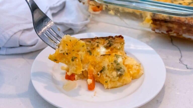 A fork holds a bite of cheesy vegetable casserole above a slice served on a white plate, with the casserole dish in the background.