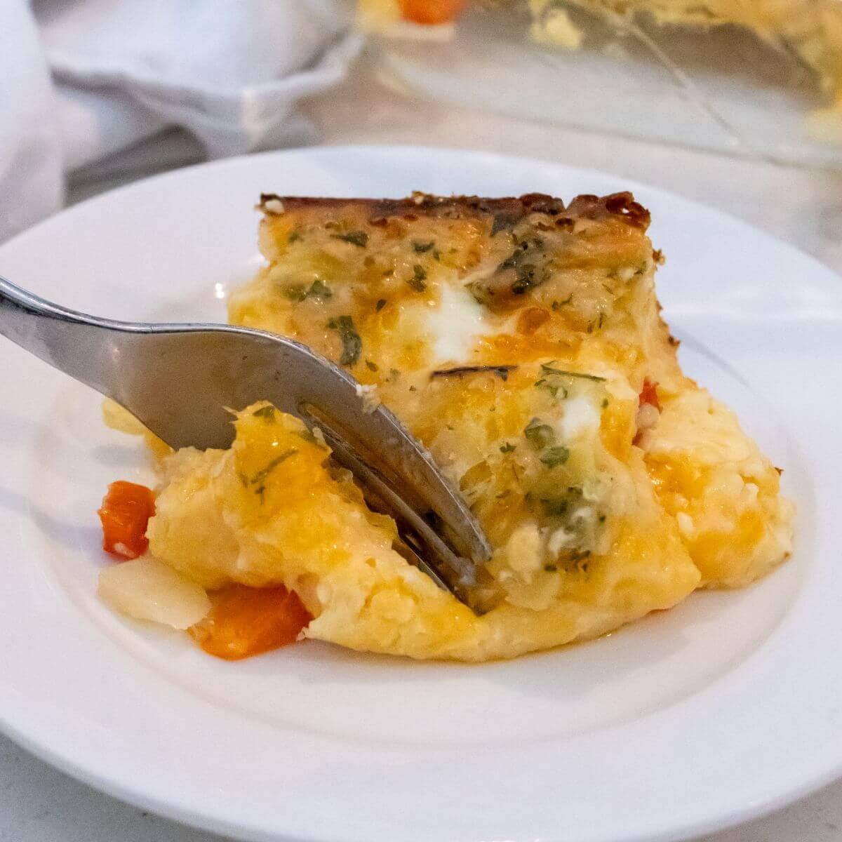 A fork cutting into a slice of cheesy egg casserole with visible red and white vegetables on a white plate.