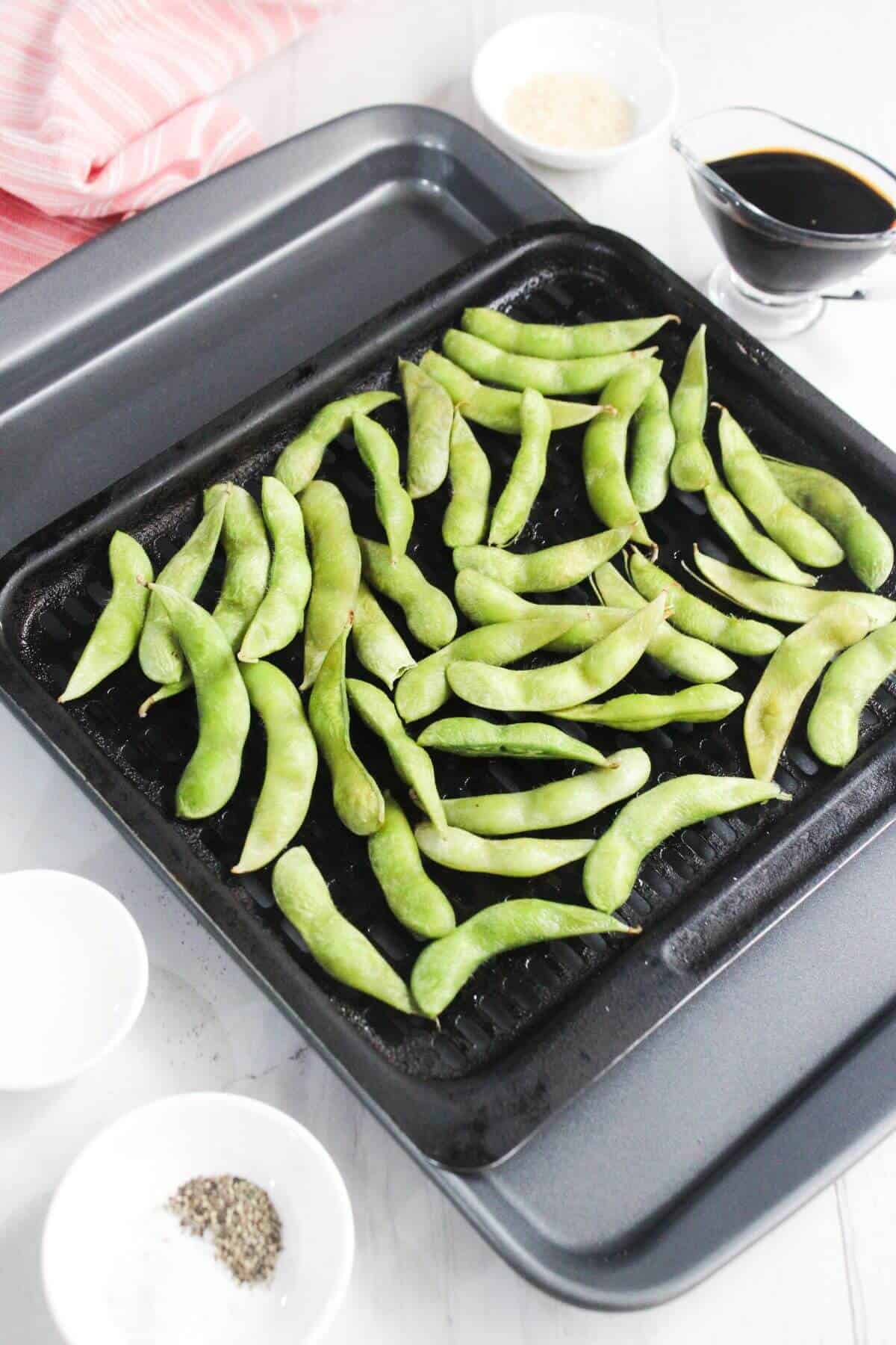 A baking tray with raw edamame pods is surrounded by small bowls containing soy sauce, sesame seeds, salt, and pepper on a white surface.