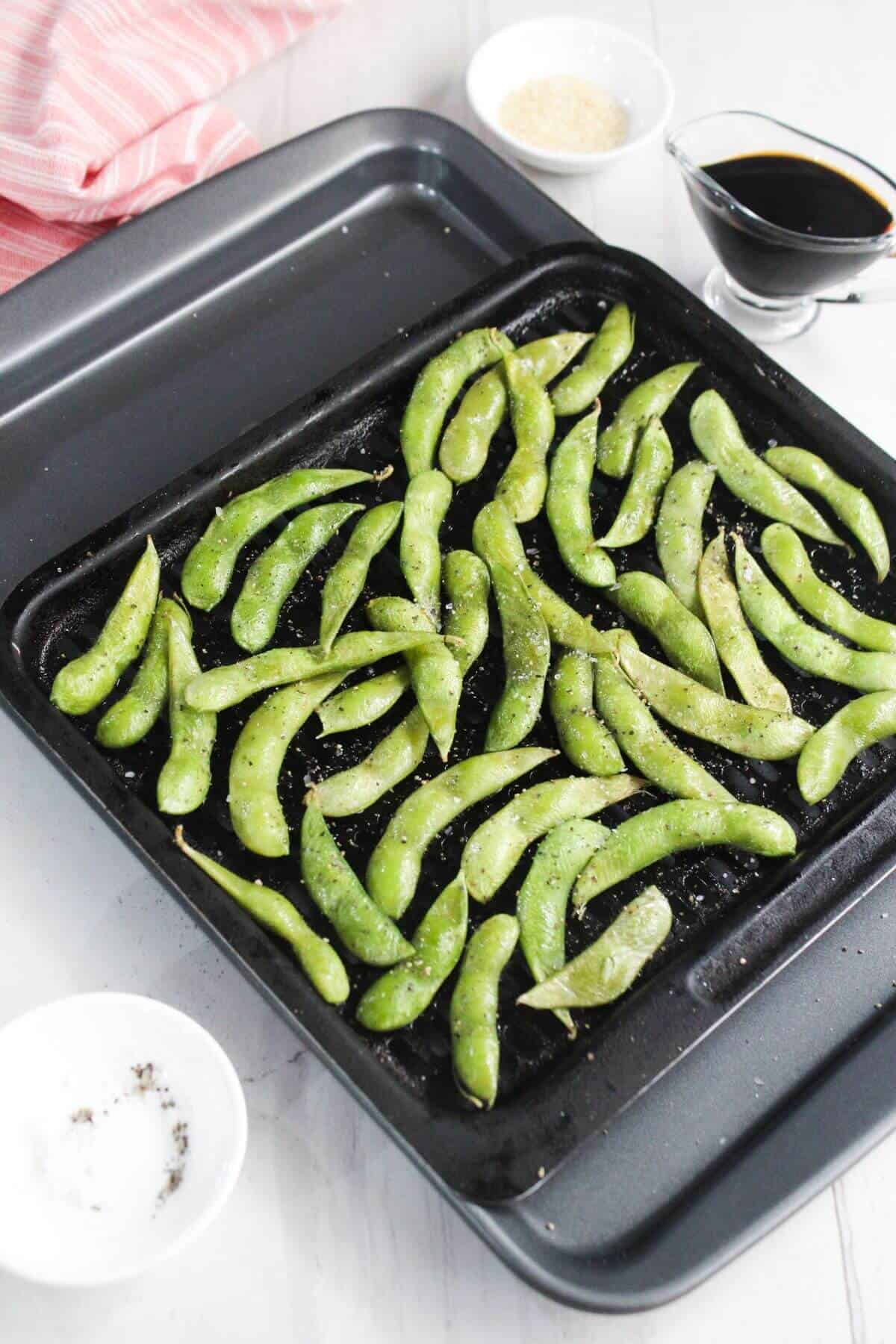 A baking tray with seasoned edamame pods, surrounded by small bowls of sesame seeds, ground pepper, and soy sauce on a white surface.