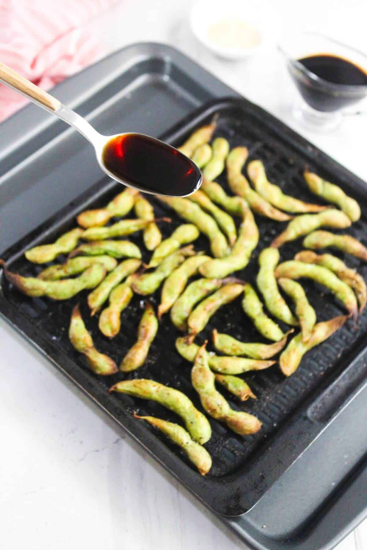 A spoon drizzling soy sauce over a tray of roasted edamame pods on a black baking sheet, with small bowls of sesame seeds and soy sauce in the background.