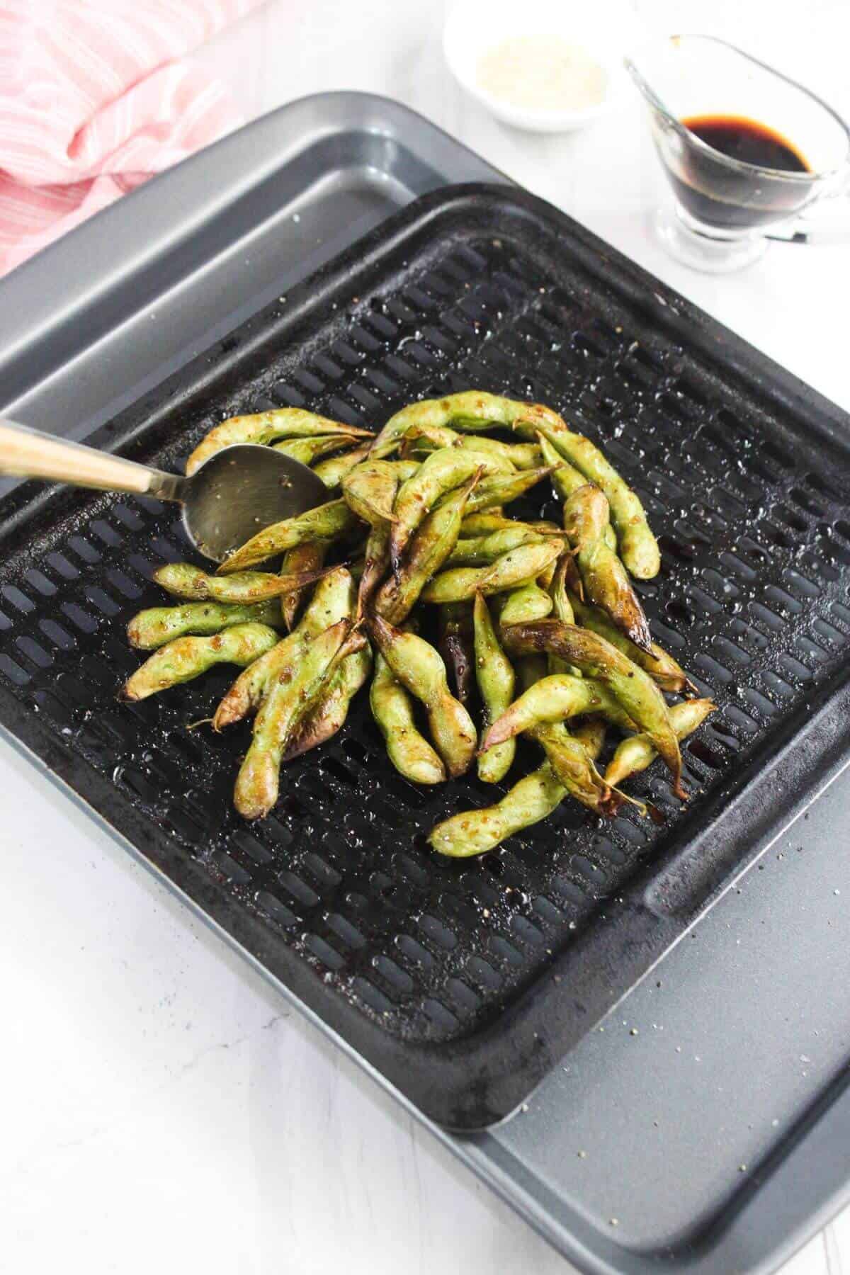 Grilled edamame pods on a grill pan with a spoon, with a bowl of dipping sauce and a bowl of sesame seeds in the background.