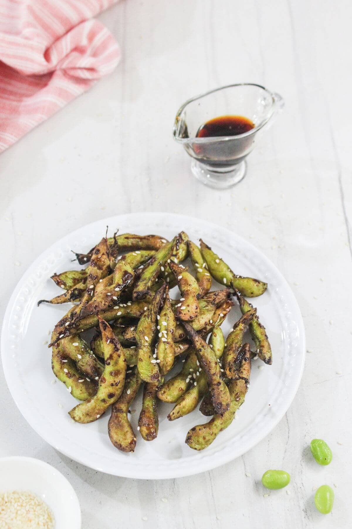 A white plate of charred edamame pods topped with sesame seeds, with a small glass container of soy sauce and a pink napkin in the background.