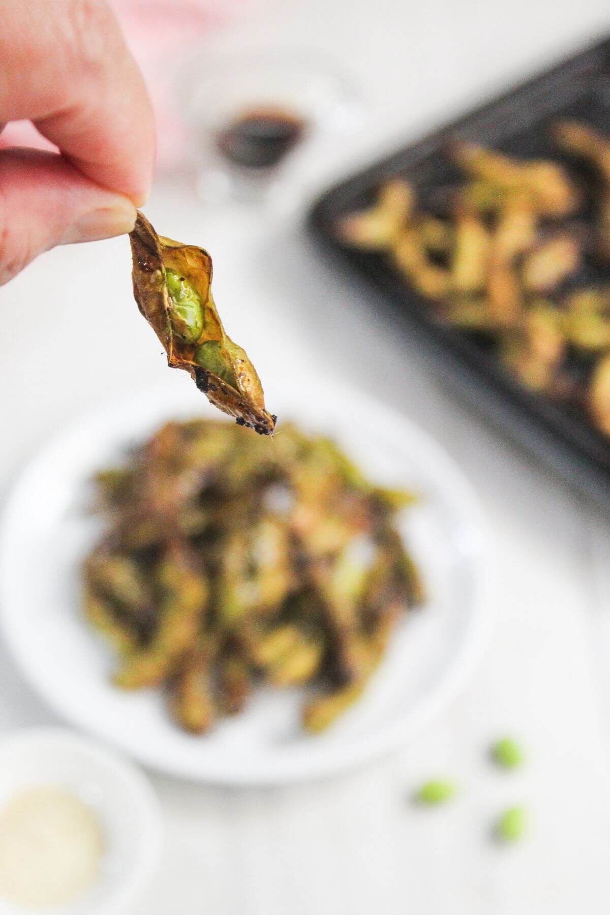 A hand holds a cooked edamame pod with a single bean exposed; a plate of additional edamame pods and a tray are blurred in the background.