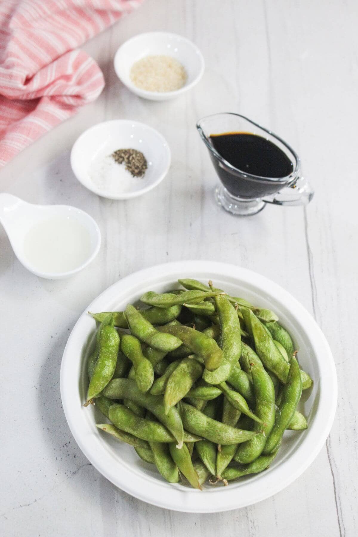 A white bowl of edamame pods is placed on a table next to small bowls of salt, pepper, sesame seeds, vinegar, and a glass container of soy sauce.