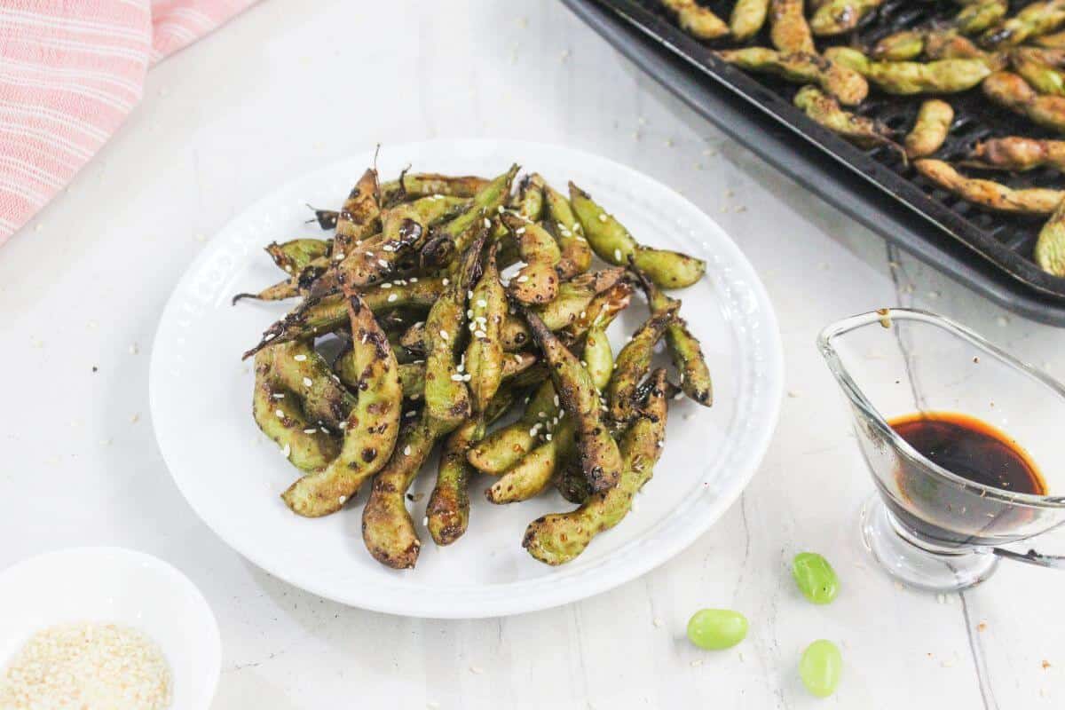 A white plate of roasted edamame pods sprinkled with sesame seeds, with a tray of more edamame and a small pitcher of soy sauce nearby on a light surface.