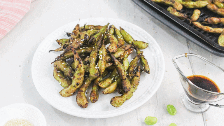 A white plate of grilled edamame sprinkled with sesame seeds, next to a small glass container of soy sauce and a tray of more edamame in the background.