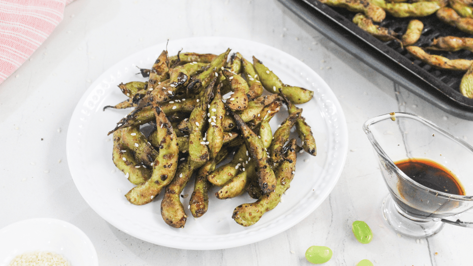 A white plate of grilled edamame sprinkled with sesame seeds, next to a small glass container of soy sauce and a tray of more edamame in the background.