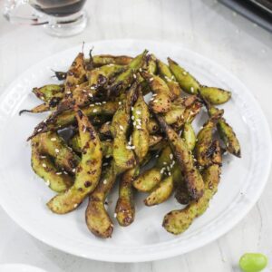 A white plate filled with grilled edamame pods sprinkled with sesame seeds, on a light-colored surface.