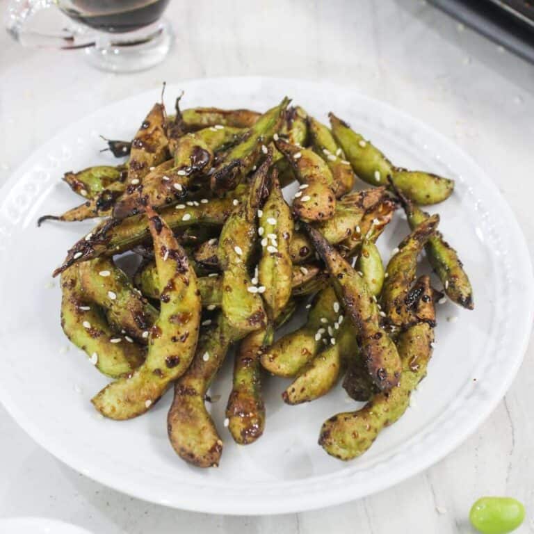 A white plate filled with grilled edamame pods sprinkled with sesame seeds, on a light-colored surface.