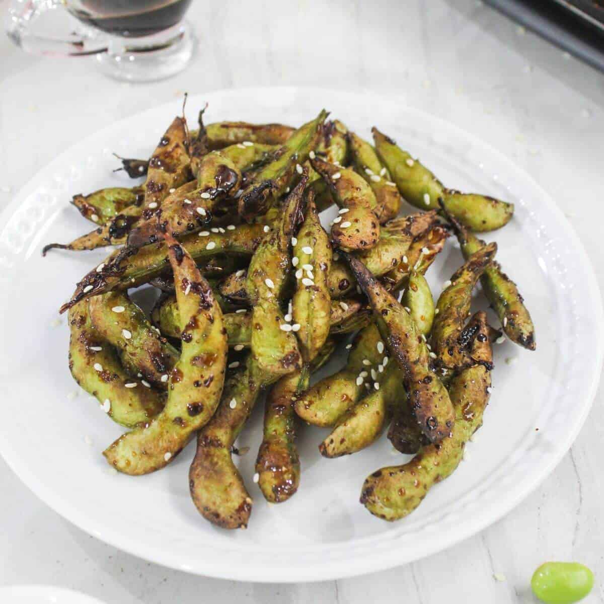 A white plate filled with grilled edamame pods sprinkled with sesame seeds, on a light-colored surface.