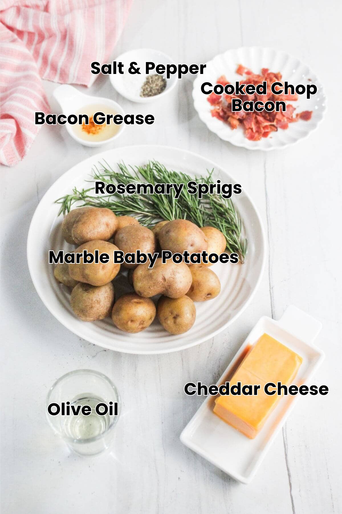 A plate of small potatoes and fresh rosemary, a block of cheddar cheese, a dish of cooked bacon pieces, a bowl of seasoning, and a glass of liquid on a white surface.