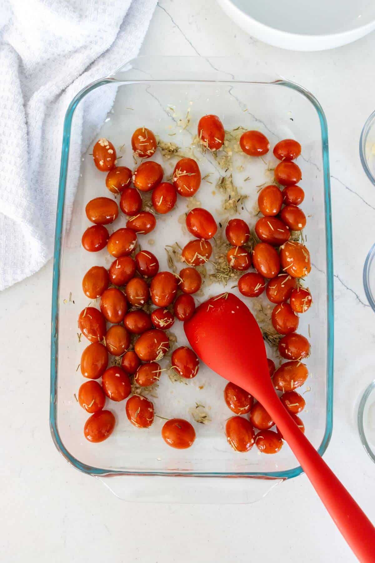 Glass baking dish with cherry tomatoes, herbs, and oil mixed with a red silicone spoon on a white countertop.