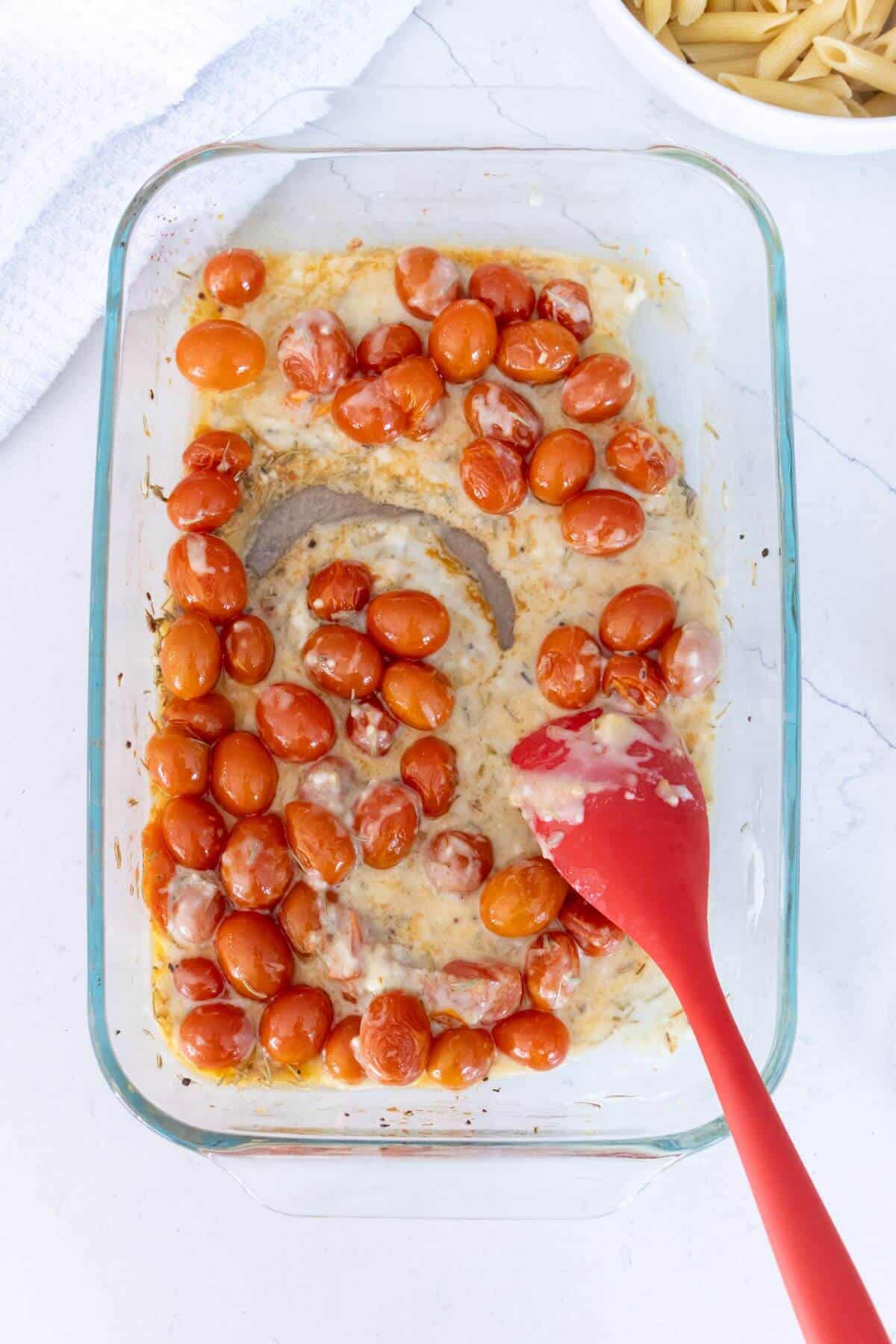 Glass baking dish with roasted cherry tomatoes and melted cheese, being stirred with a red spatula on a white surface.
