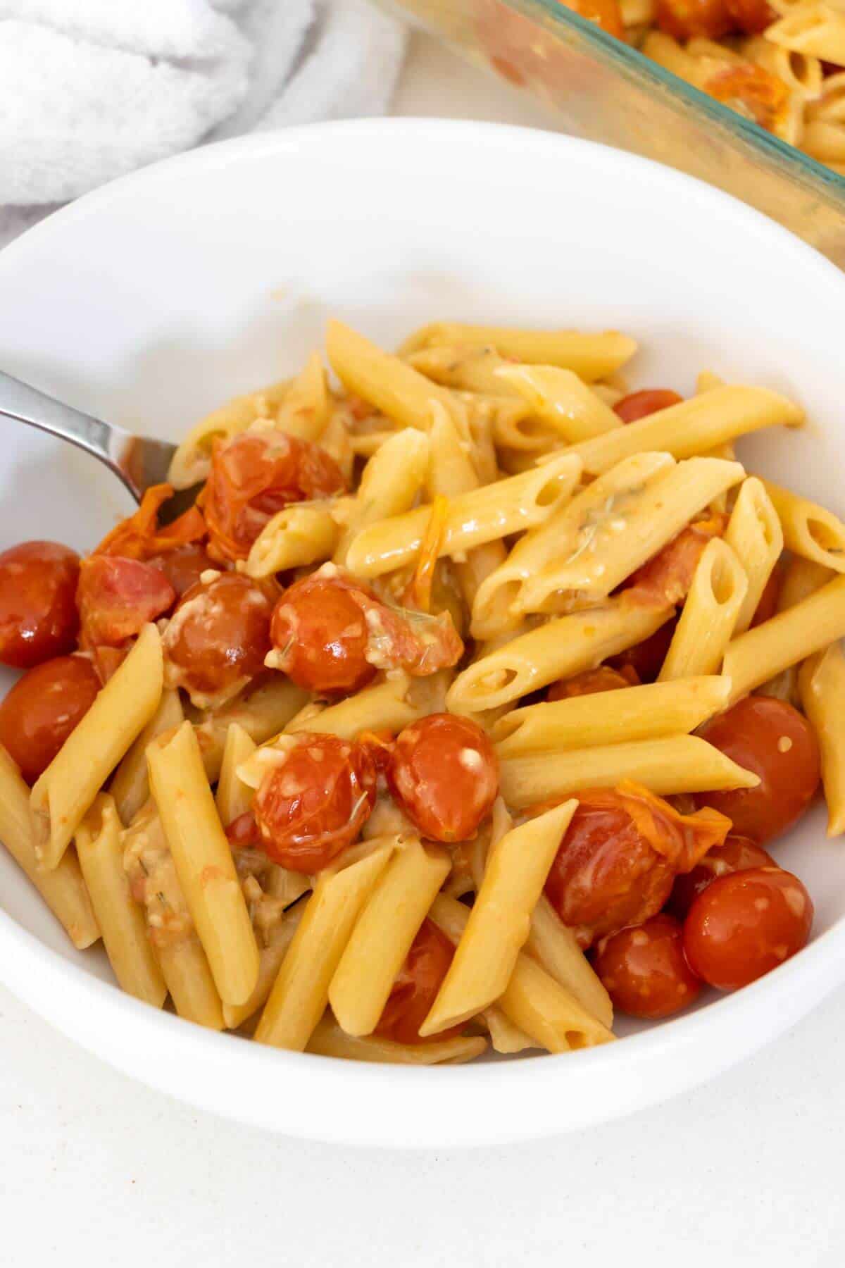 A white bowl filled with penne pasta, whole cherry tomatoes, and a creamy sauce, with a fork resting on the side of the bowl.
