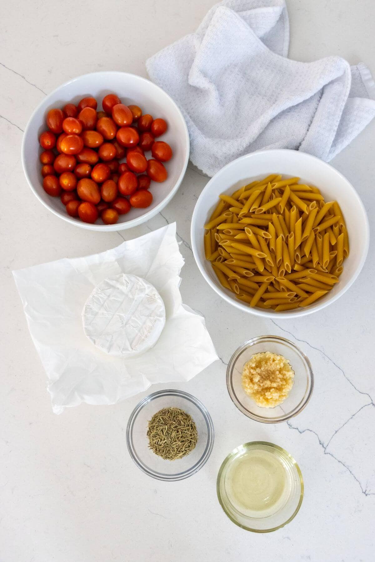 Bowls of grape tomatoes, uncooked penne pasta, a round of brie cheese, dried herbs, minced garlic, and oil on a white countertop with a folded kitchen towel.