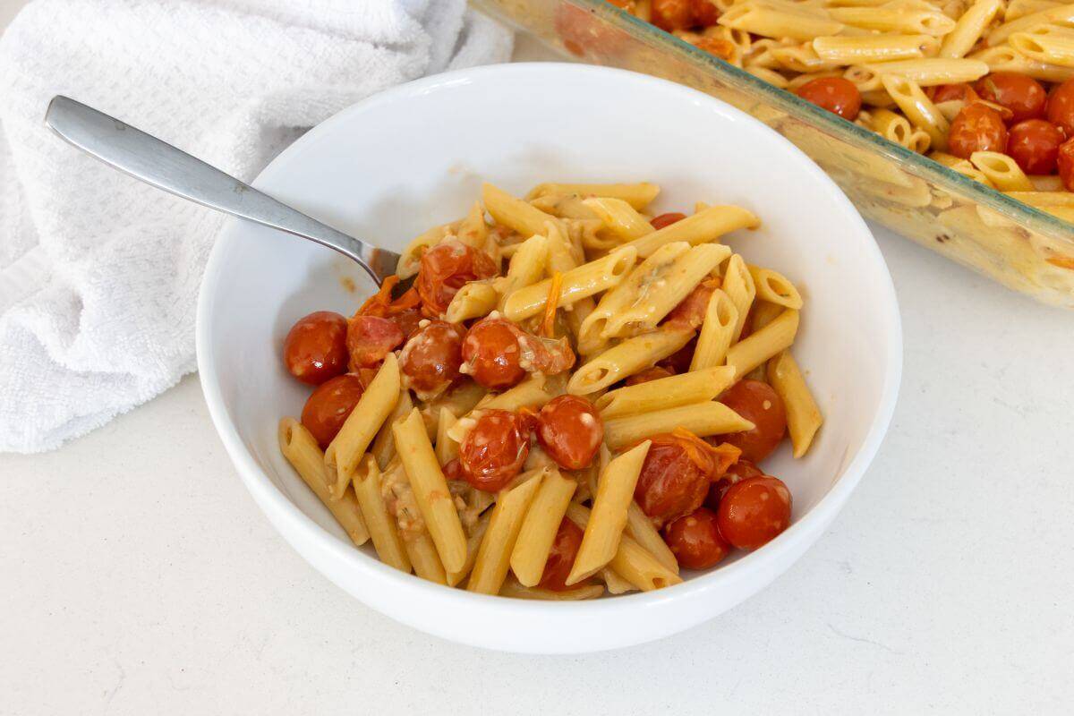 A white bowl filled with penne pasta, cherry tomatoes, and creamy sauce, with a fork inside. A white towel and a baking dish of pasta are in the background.