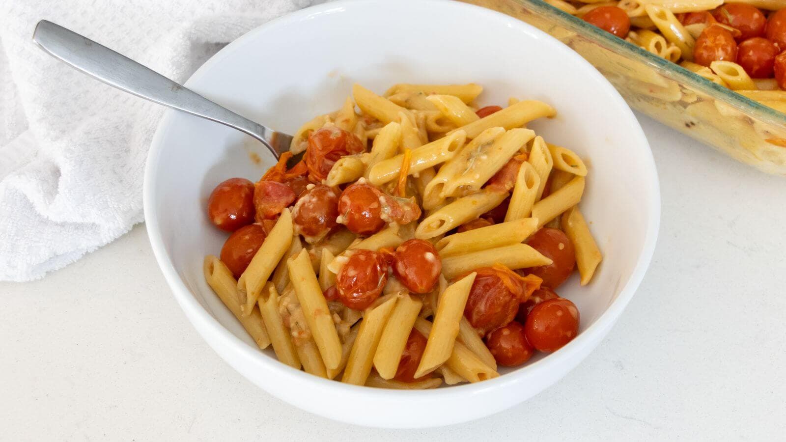 A white bowl filled with penne pasta, cherry tomatoes, and a creamy sauce, with a fork in the bowl. A glass baking dish and a white towel are nearby.
