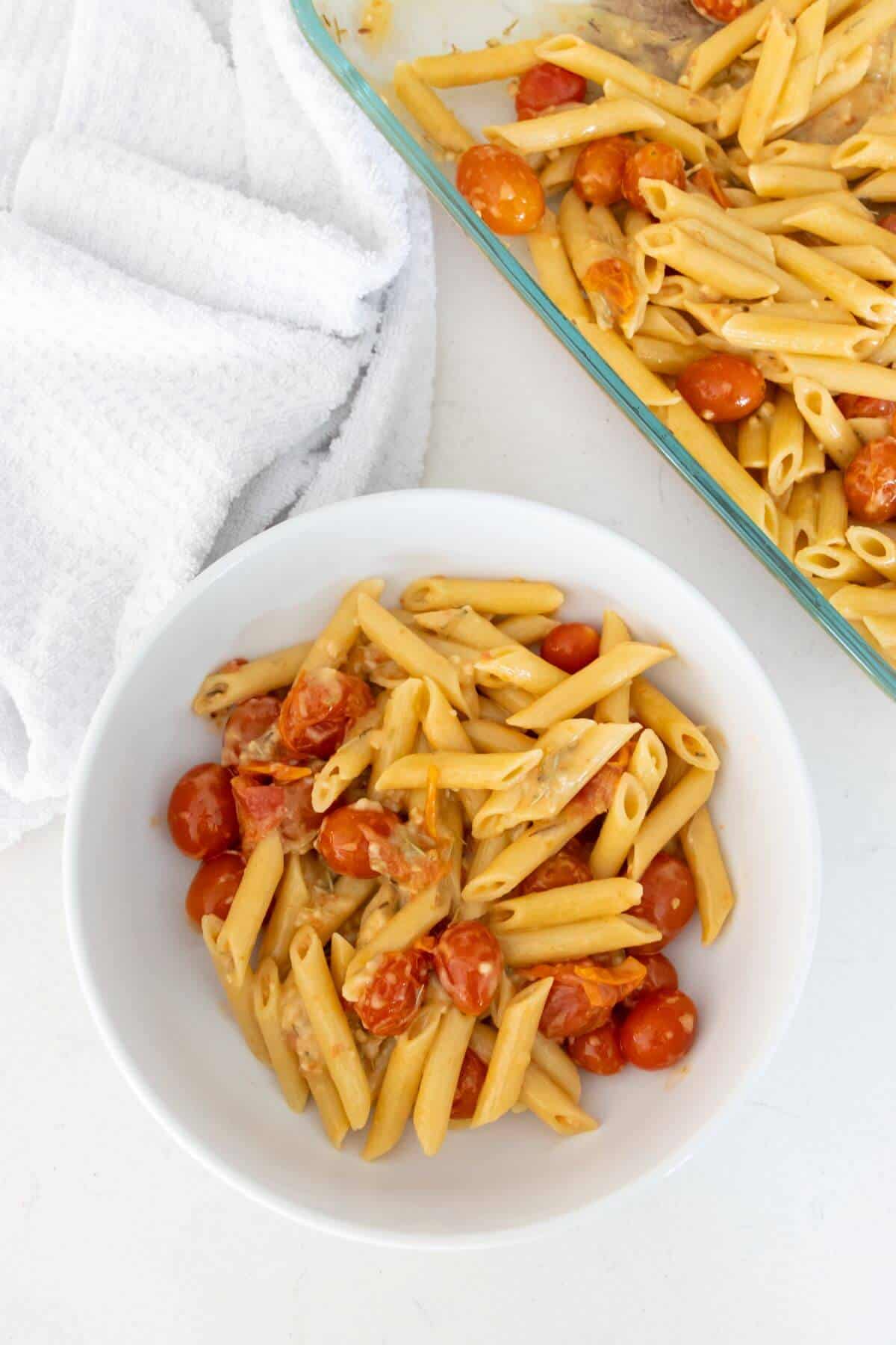 A white bowl of penne pasta with cherry tomatoes and sauce, next to a baking dish with more pasta. A white towel is placed nearby.