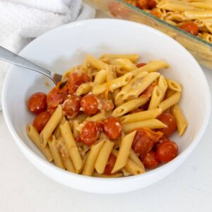 A white bowl filled with penne pasta, cherry tomatoes, and a creamy sauce, with a fork resting inside. A baking dish with more pasta is visible in the background.
