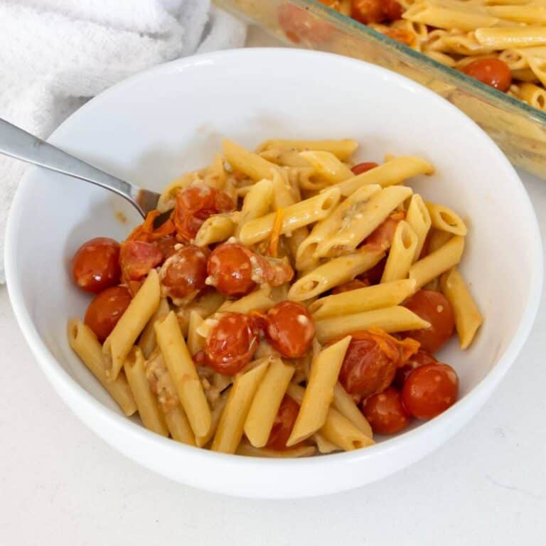 A white bowl filled with penne pasta, cherry tomatoes, and a creamy sauce, with a fork resting inside. A baking dish with more pasta is visible in the background.
