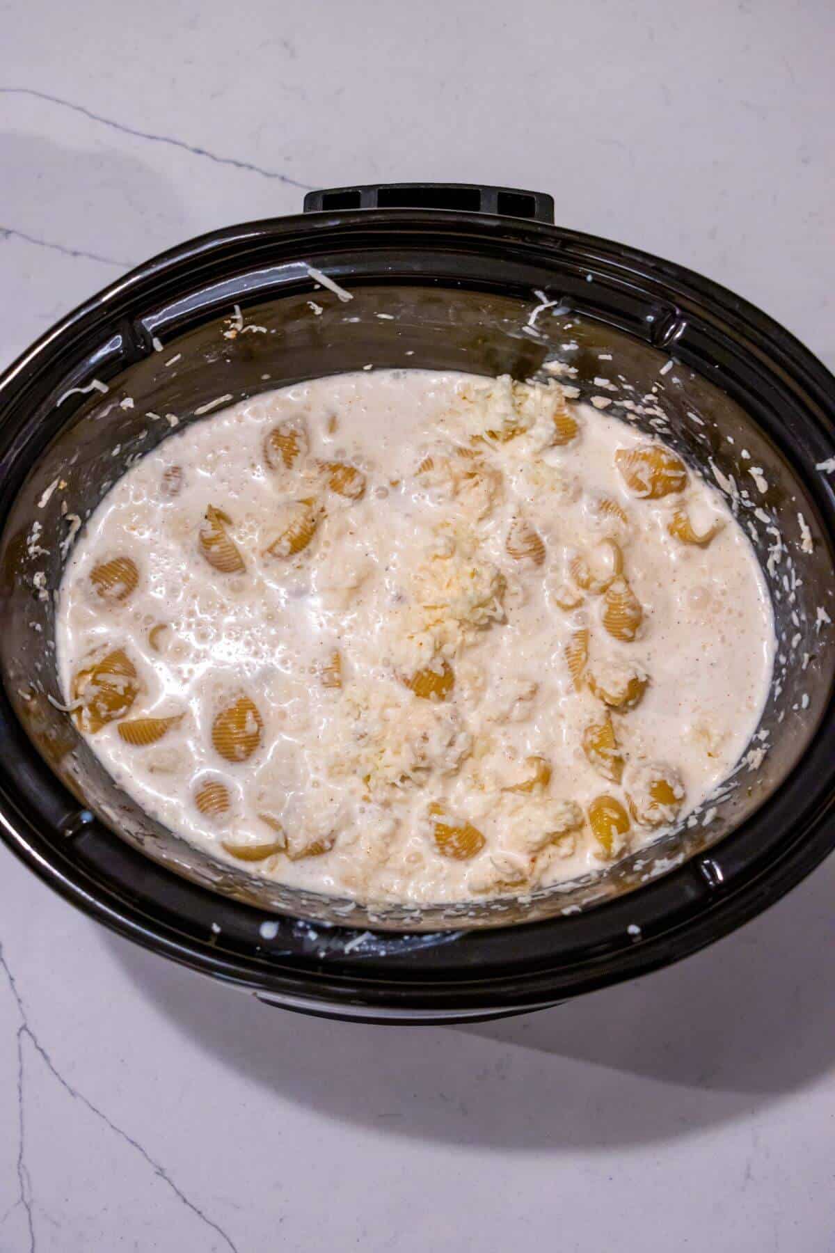 A slow cooker filled with a creamy mixture of pasta shells, cheese, and sauce on a light countertop.