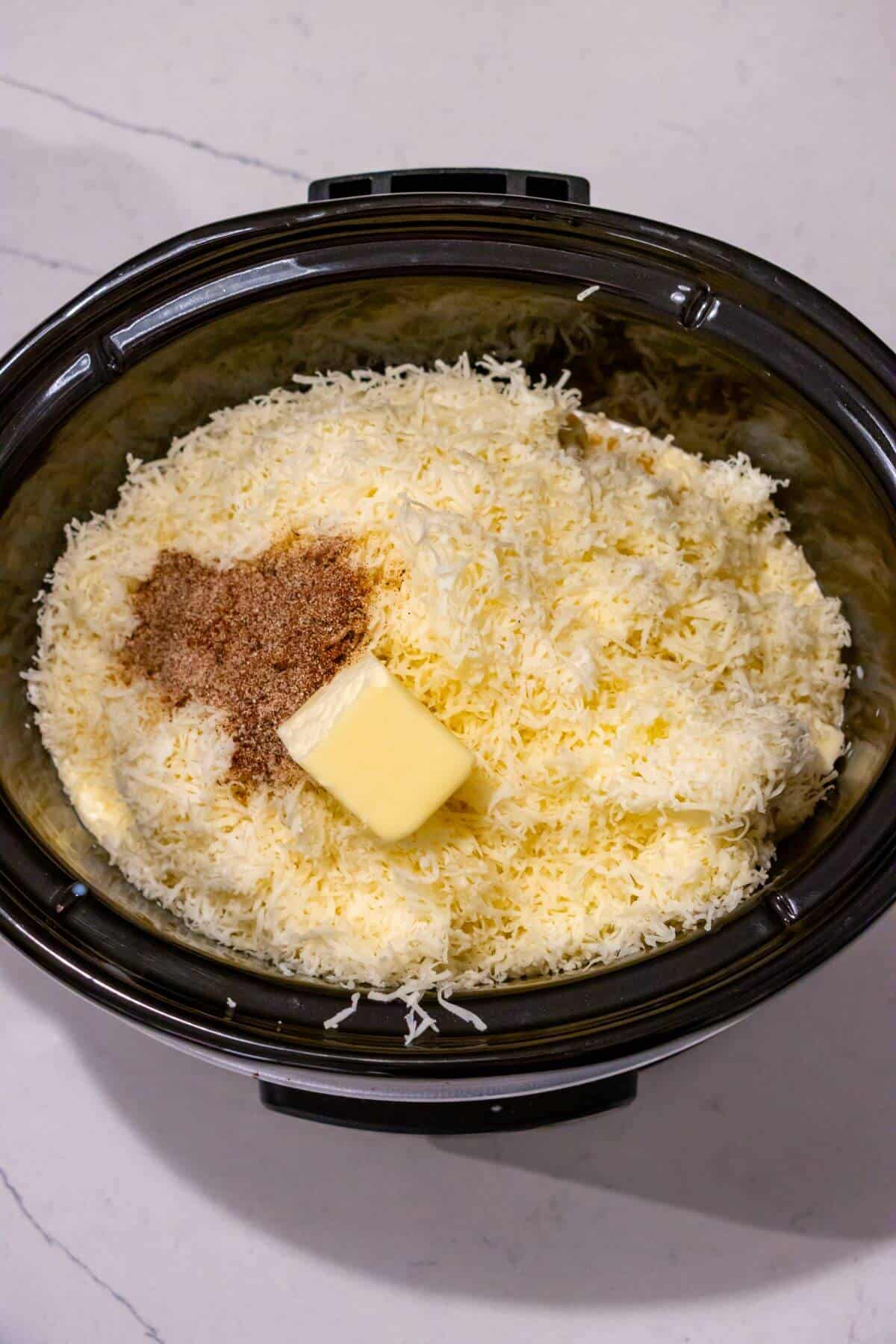 Shredded cheese, butter, and seasoning are placed in a slow cooker on a light-colored countertop.