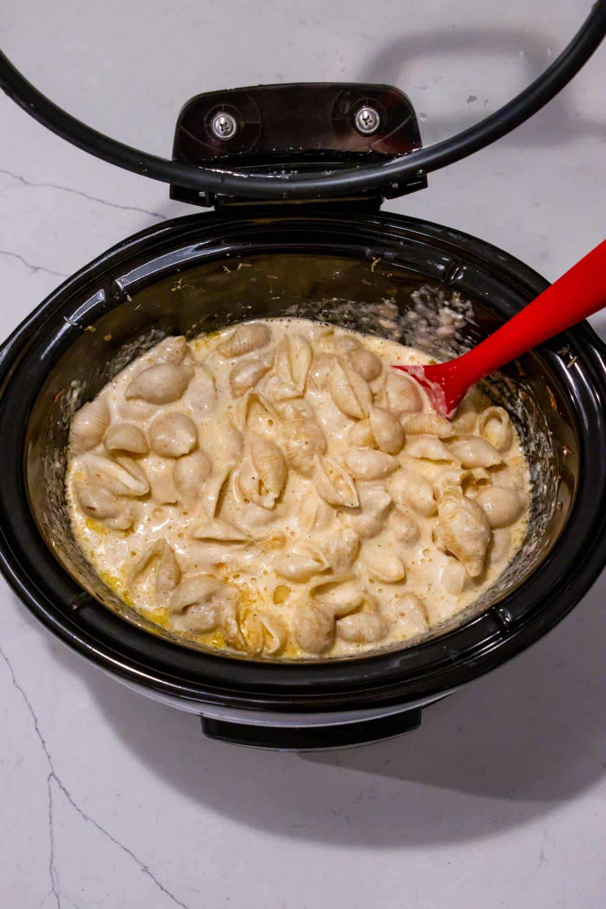 Cooked pasta shells in a creamy sauce inside a slow cooker, with a red spatula resting in the mixture.