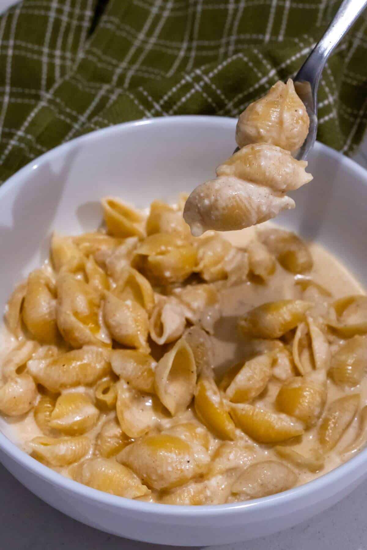 A white bowl filled with creamy macaroni and cheese shells, with a spoon lifting a portion. A green plaid cloth is in the background.