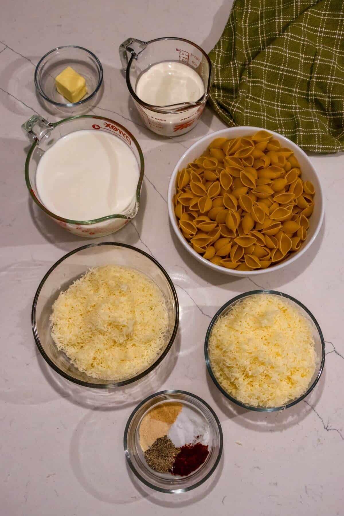 Ingredients for a pasta dish arranged on a counter: pasta shells, shredded cheese, milk, cream, butter, and various spices in small bowls. A green checked cloth is nearby.