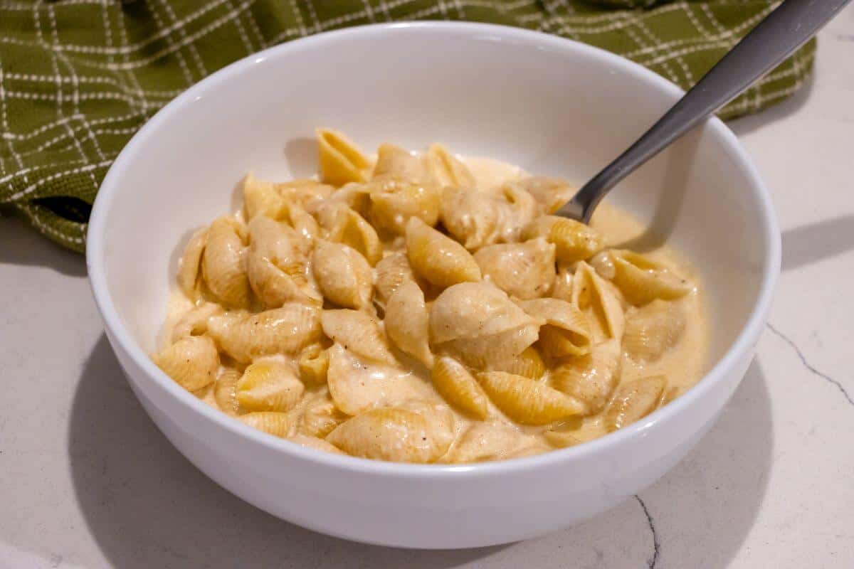 A white bowl filled with creamy macaroni and cheese pasta shells, with a spoon resting inside, sits on a countertop next to a green checkered cloth.