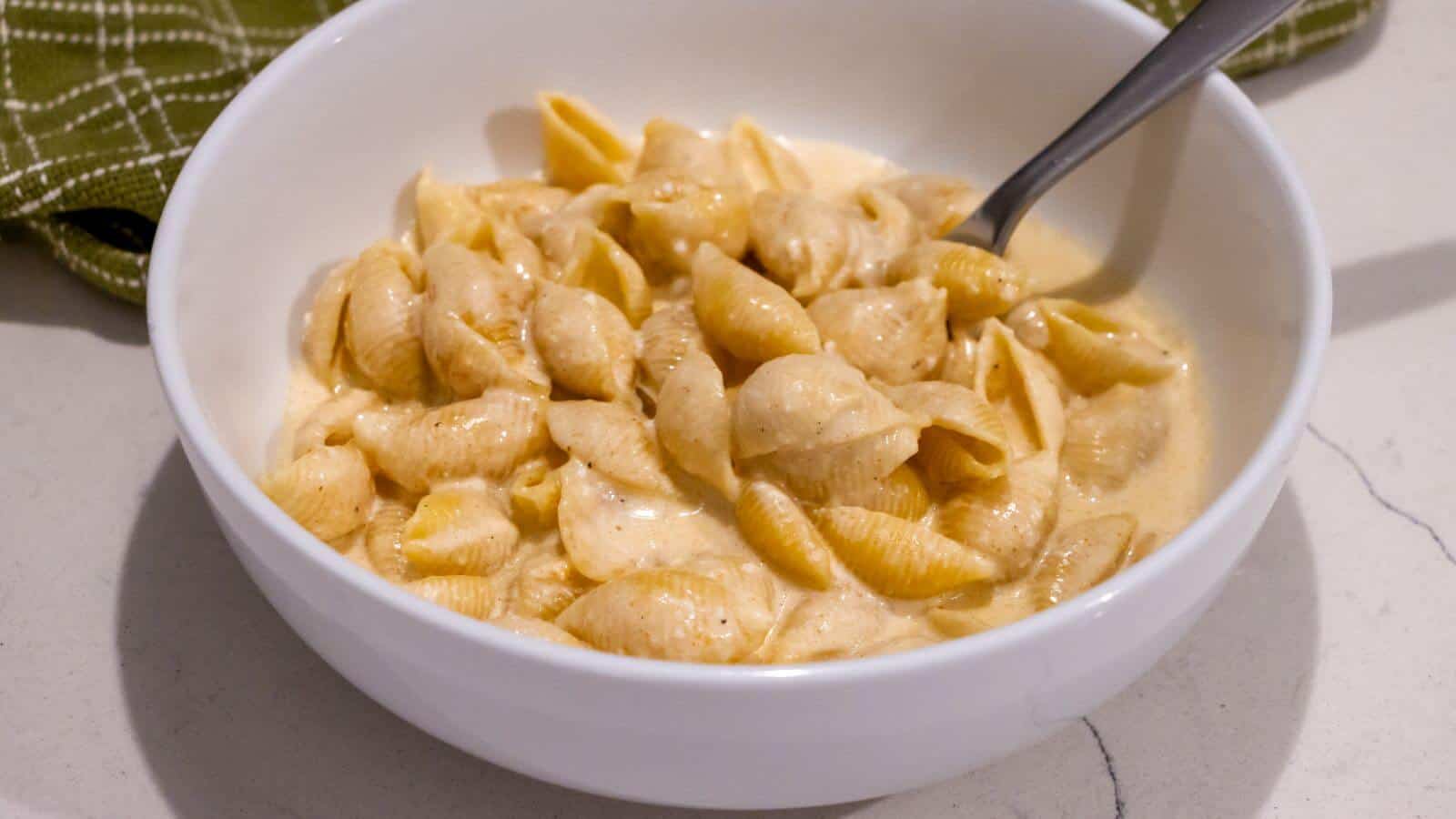 A white bowl filled with shell pasta coated in creamy cheese sauce, with a metal spoon inside, placed on a light countertop.
