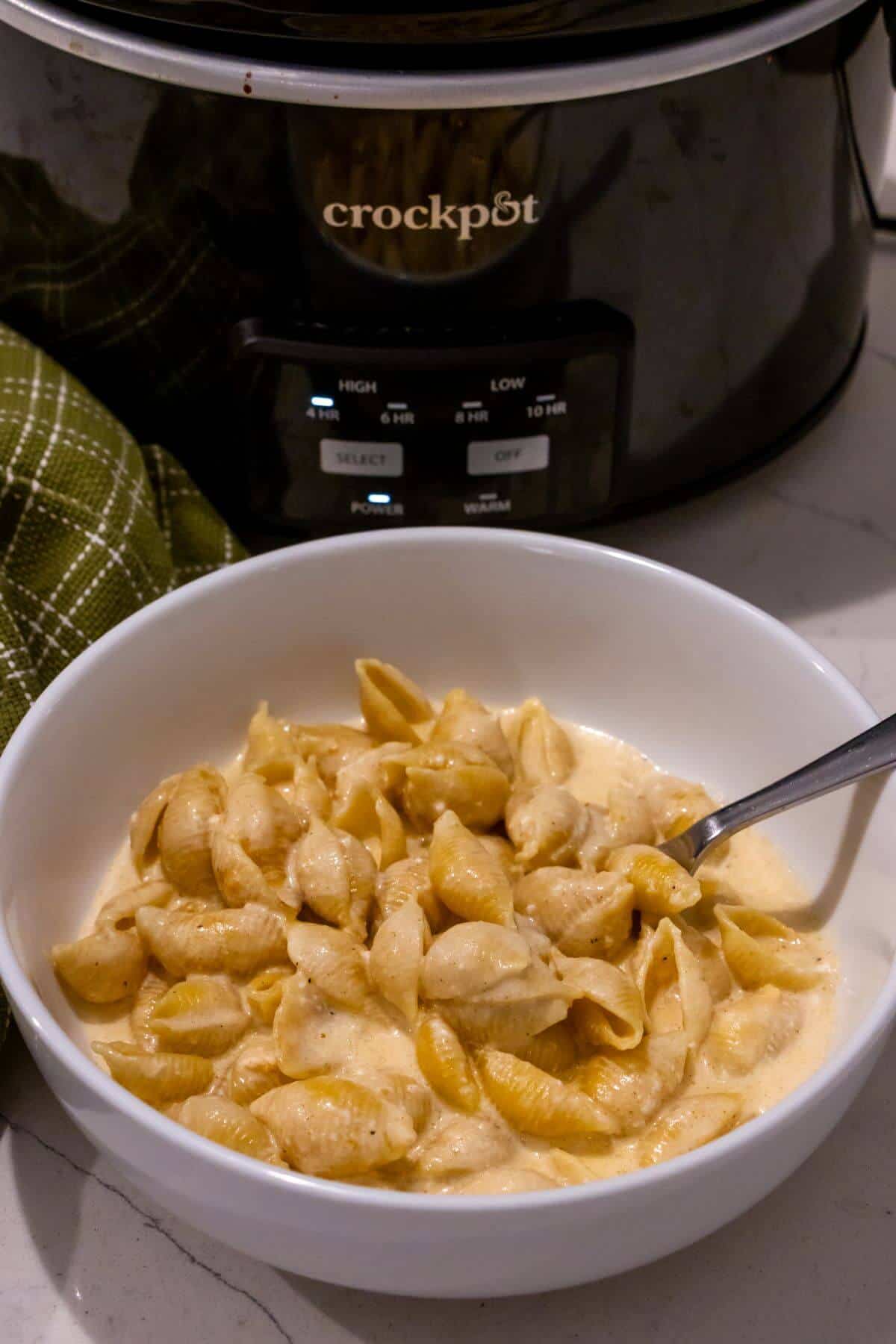 A white bowl filled with creamy macaroni and cheese sits in front of a black Crockpot on a kitchen counter, with a green cloth nearby.
