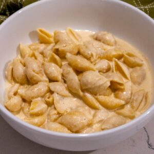 A white bowl filled with pasta shells coated in a creamy cheese sauce, placed on a light-colored surface with a green cloth in the background.