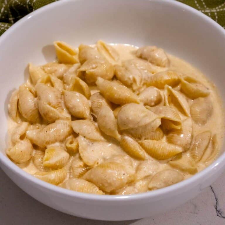 A white bowl filled with pasta shells coated in a creamy cheese sauce, placed on a light-colored surface with a green cloth in the background.