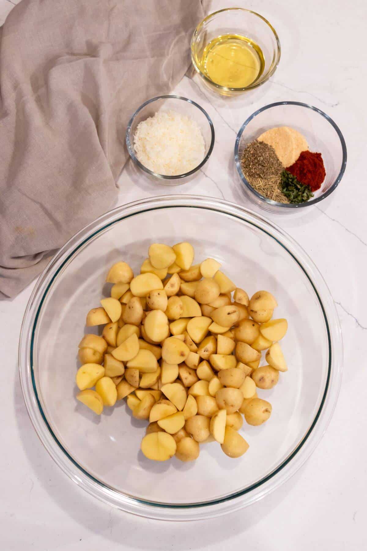 A glass bowl with quartered baby potatoes, next to bowls of olive oil, grated cheese, and assorted seasonings on a white surface with a beige cloth.
